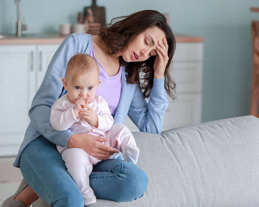 a mother holding her baby in the living room, her hand is on her forehead.